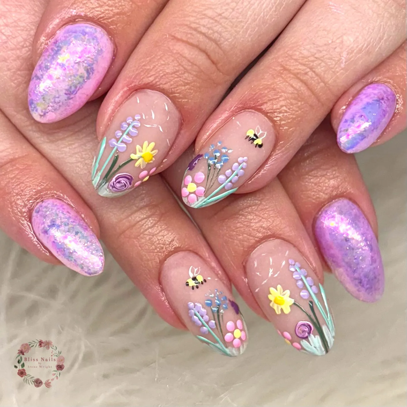 These medium-length, almond-shaped nails mix soft lavender and lilac shimmer with delicate hand-painted floral details on a sheer nude base. The accent nails feature tiny wildflowers in pastel yellow, pink, blue, and purple, slender green stems, and miniature bees hovering between the blooms, while the surrounding nails glow with a mottled, watercolor-style purple shimmer that catches the light. The overall look feels organic, dreamy, and slightly untamed, leaning into the wilderkind aesthetic with its nature-led details and soft, storybook charm. This design is perfect for spring, garden parties, or anyone drawn to whimsical, nature-inspired nail art.
