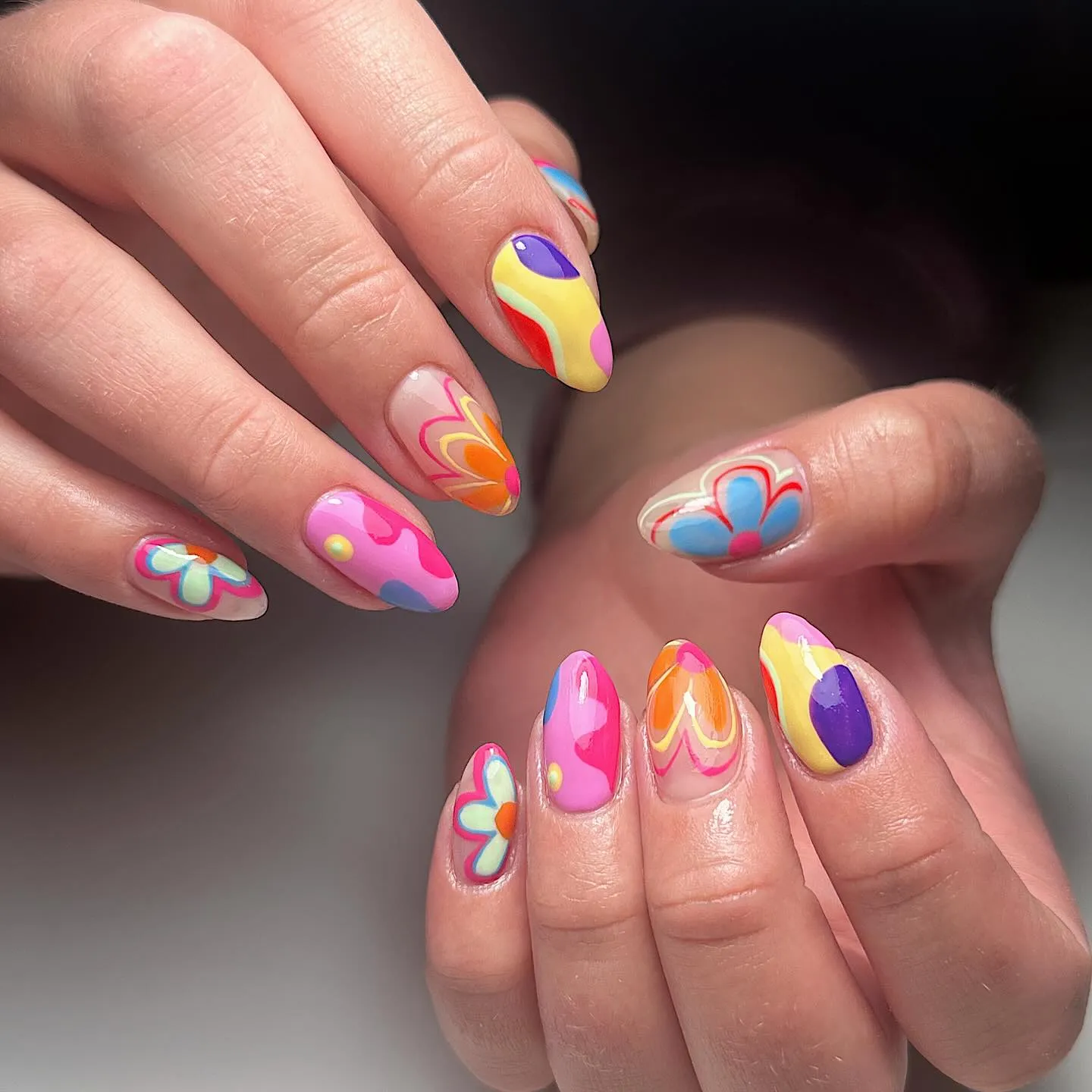 These long, almond-shaped nails feature bright pink, orange, yellow, blue, and purple colours. Each nail has a different design, including bold curved stripes, abstract swirls, heart shapes, and retro-style flowers, all finished with a glossy shine. The overall aesthetic feels playful and nostalgic, with strong 60s and 70s retro vibes and a fun, mismatched look across both hands. This vibrant manicure is perfect for summer, holidays, or festivals when you want colourful statement nails that stand out.