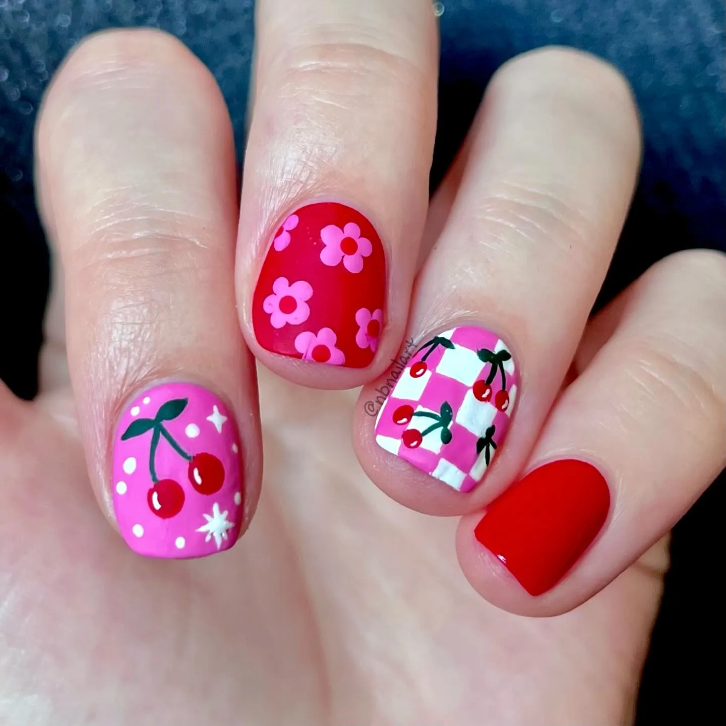 These short, square-shaped nails feature a playful mix of bright red and bubblegum pink, with bold accent designs on each nail. One nail shows sweet cherry artwork on a pink base with white polka dots, another has a red background scattered with simple pink flower shapes, and a third combines a pink-and-white gingham pattern topped with tiny cherries. The solid red nails ground the look, while the illustrated details give it a cute, mid century retro-inspired aesthetic that feels very vintage picnic meets pop art. This design is perfect for summer, Valentine’s season, or anytime you’re leaning into fun, cheerful nail art.