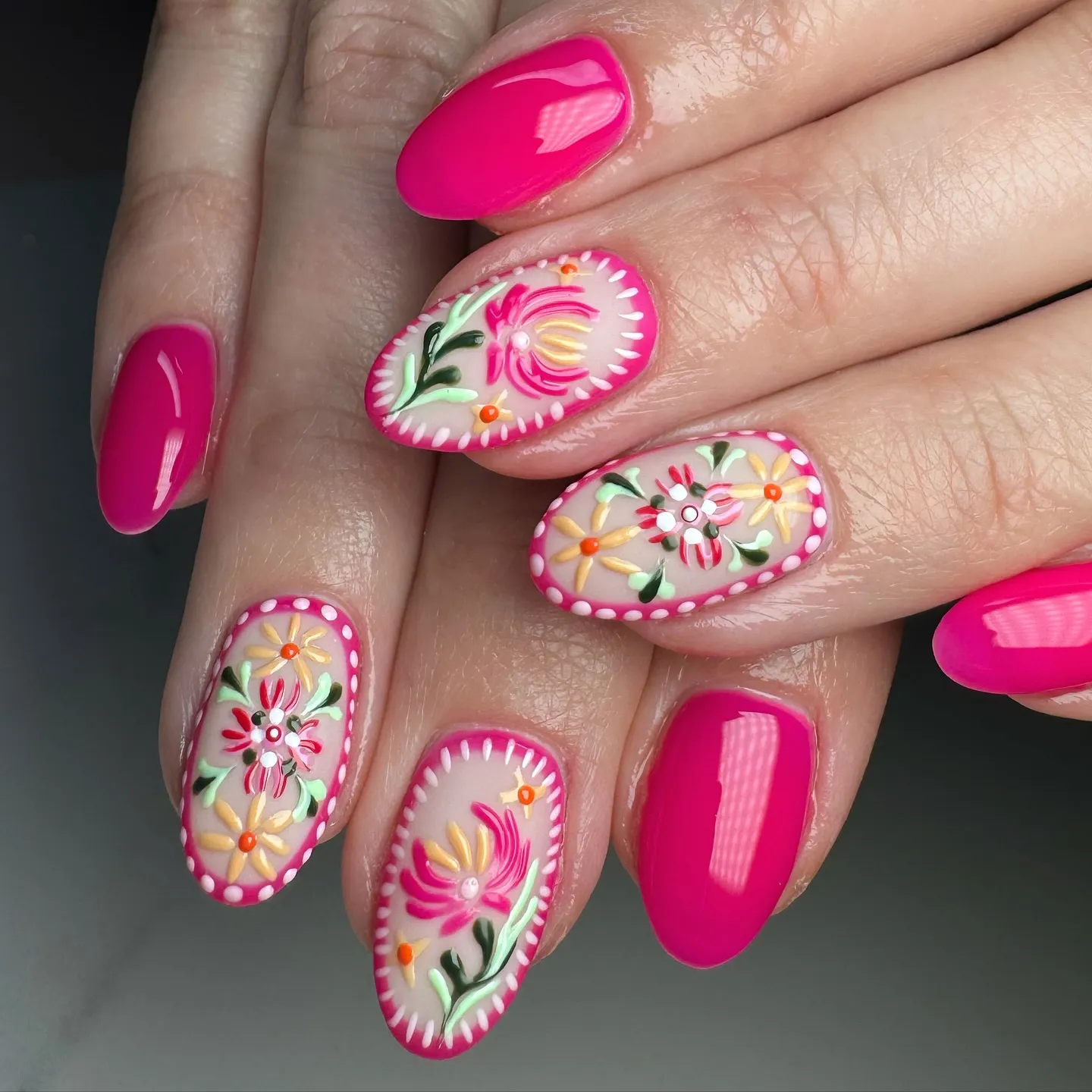 These medium-length, oval-shaped nails feature a bright hot pink base paired with detailed floral nail art. The accent nails are painted with hand-drawn flowers in soft yellow, coral, white, and green, set against a sheer nude background and framed with tiny dotted borders for a folk-art feel. The raised floral details give the design texture, while the solid pink nails keep the look bold and balanced. This set feels cheerful and artistic, perfect for spring, summer, or anyone who loves colourful nails with a handcrafted, feel-good vibe.