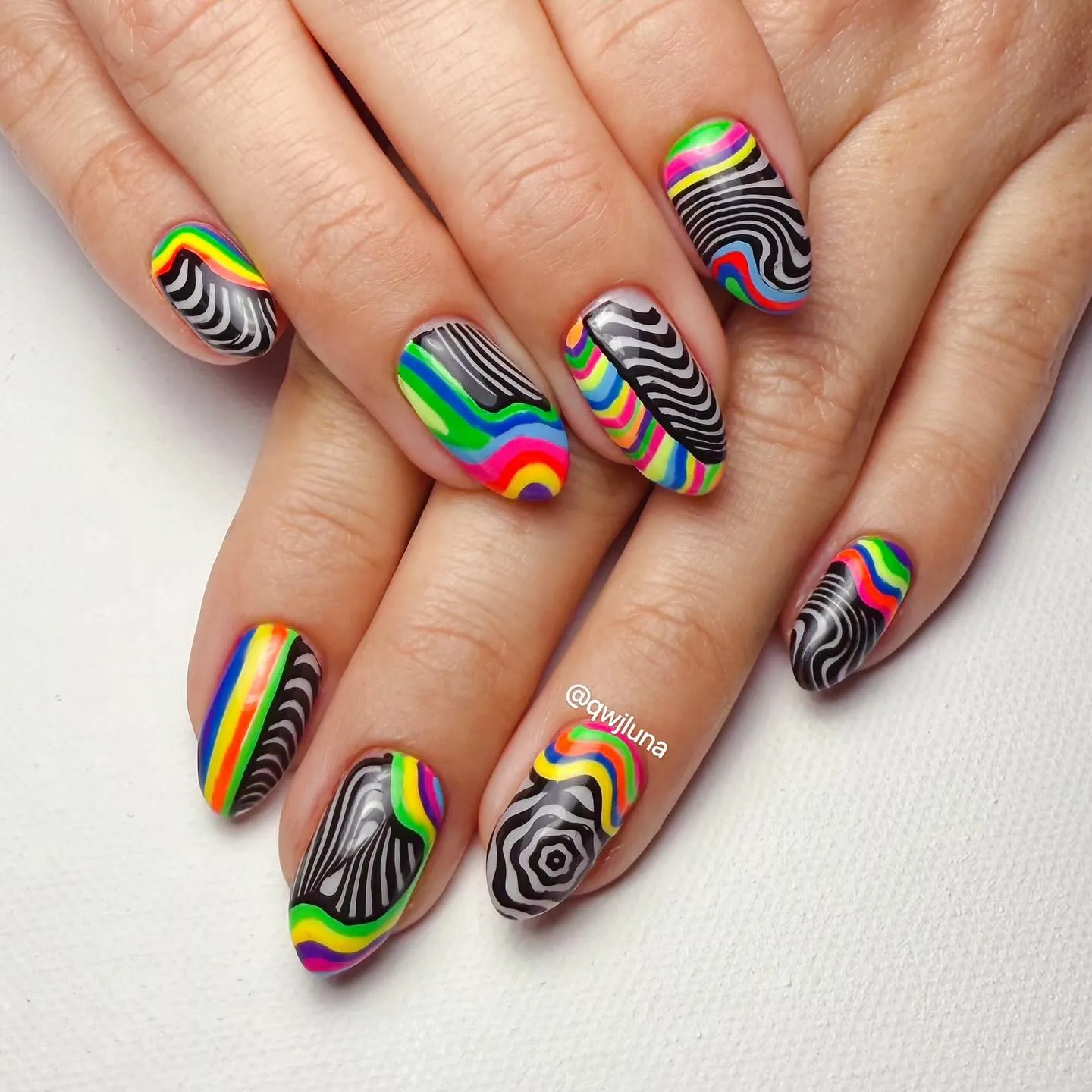 These medium-length, almond-shaped nails combine bold rainbow stripes with high-contrast black-and-white optical patterns. Bright bands of red, orange, yellow, green, blue, and pink ripple across each nail, layered alongside swirling lines, warped waves, and hypnotic concentric designs that create a striking visual illusion effect. The glossy finish makes the colours pop even more, giving the whole set a graphic, psychedelic aesthetic that feels very retro-meets-modern. This design is perfect for summer, Pride season, music festivals, or anyone who loves vibrant nails that refuse to blend in.